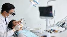 A male dentist wearing loupes and a mask examines a young male patient's open mouth in a modern dental chair, with an overhead light, monitor screen, and equipment nearby in a clinic setting
